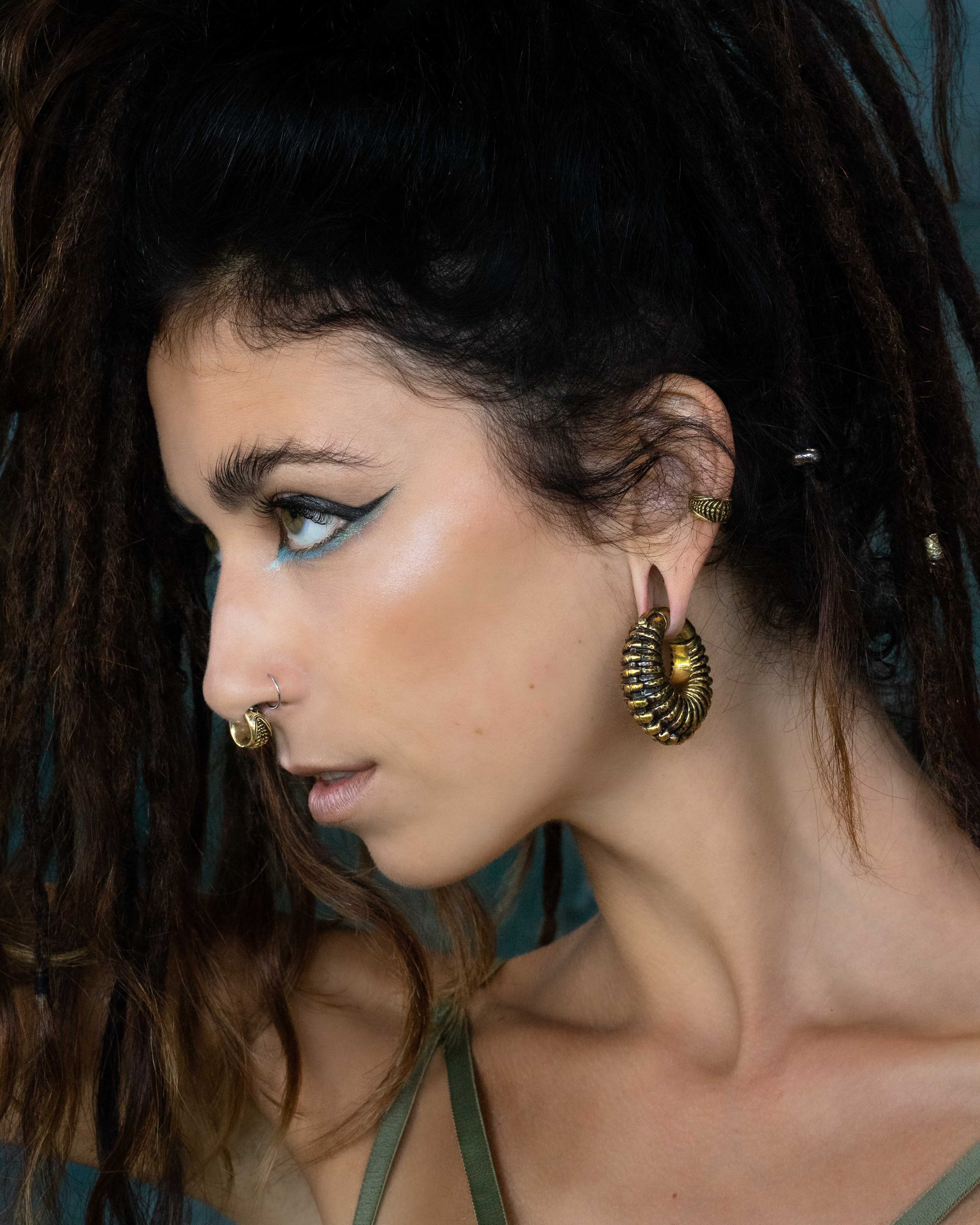 A woman with long, dark dreadlocks is shown in profile against a dark background. She wears large, ornate gold hoop earrings and a gold nose ring combined with TRILOBITE Large Biomechanical Clicker Ear Weights in Gold Brass by Kaya Movement at her 0 gauge ears. Her bold, artistic makeup features winged eyeliner, and she has a focused, intense expression.